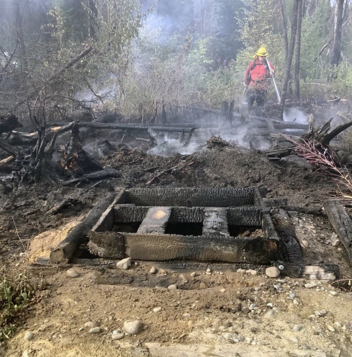 A firefighter sprays down hotspots on a wildfire behind a burned-up fish smoker.