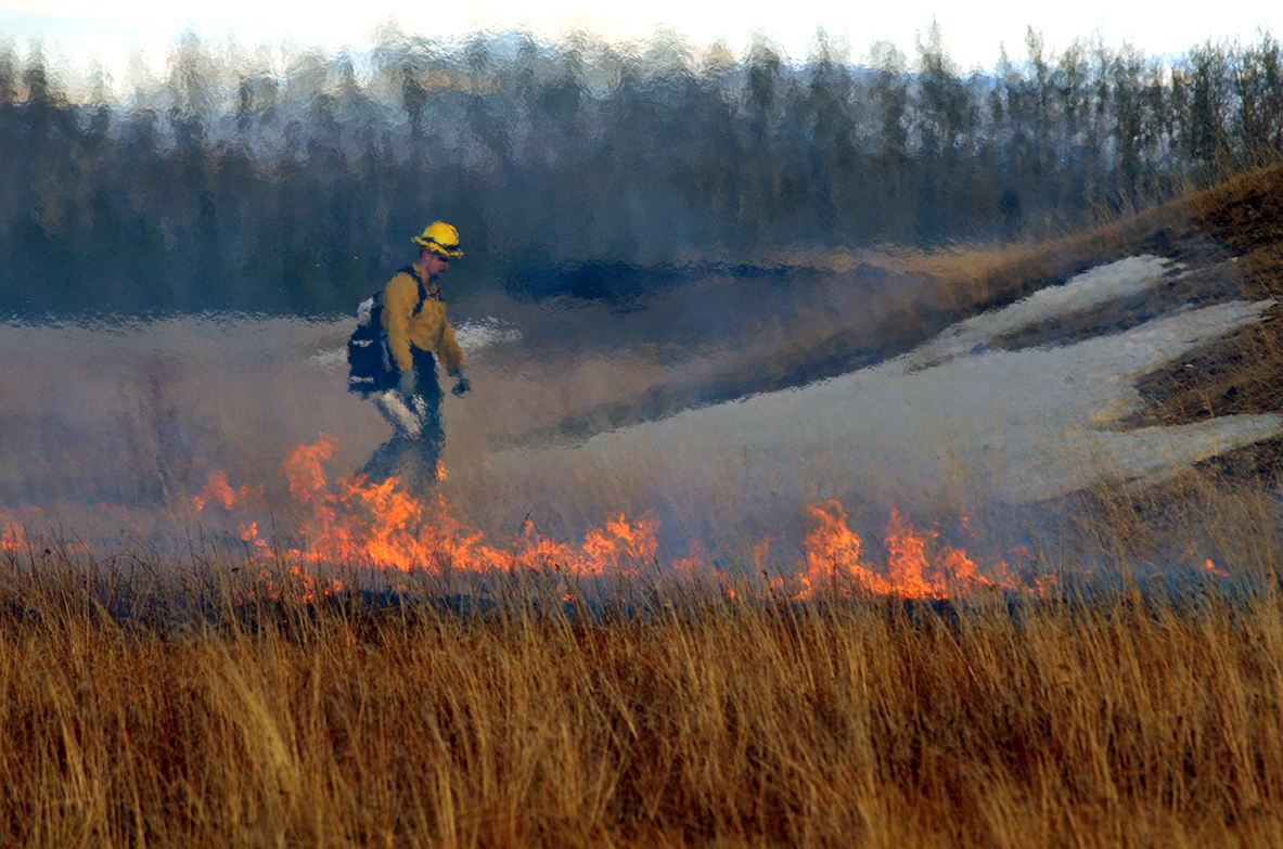 A firefighter blurred by the heat rising from grass burning at his feet.