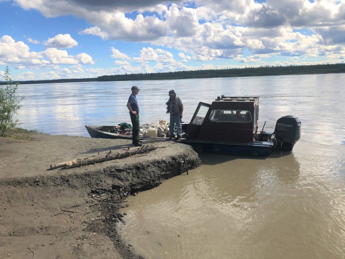 Motor boat on Tanana River with used fire hose loaded inside docked on the shore at Manley. Boat driver and firefighter talking on near boat. Clouds with blue sky and river is brownish color.