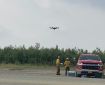 Photo of a drone in the sky with firefighters on the ground wearing yellow nomex shirts and green nomex pants. They are standing next to a red Chevrolet truck in a gravel lot. Green trees and brush in the background.